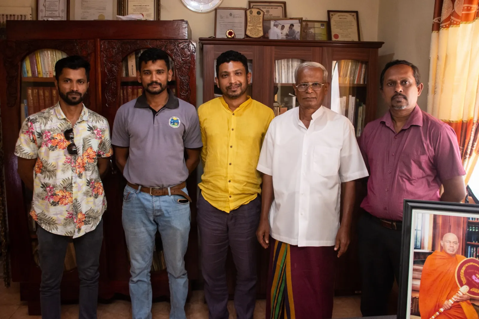 Five men standing together in an Ayurvedic doctor's study lined with bookshelves and framed certificates