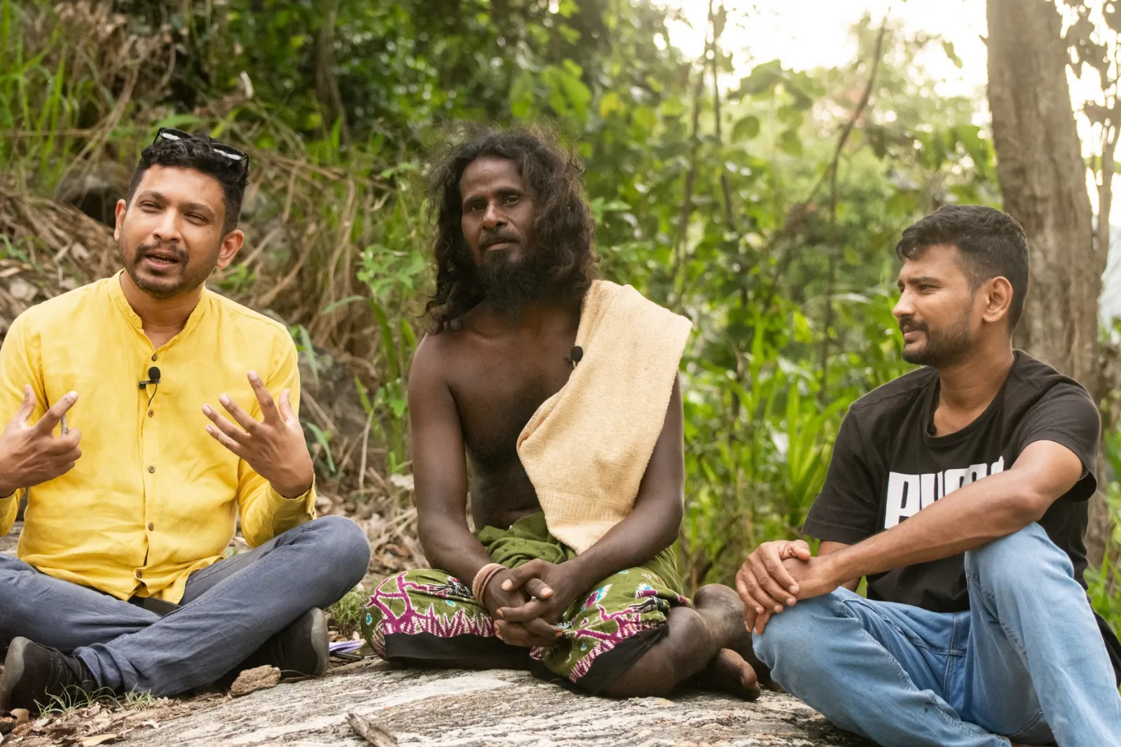 Three men in conversation, seated cross-legged on a rock in a Sri Lankan forest clearing