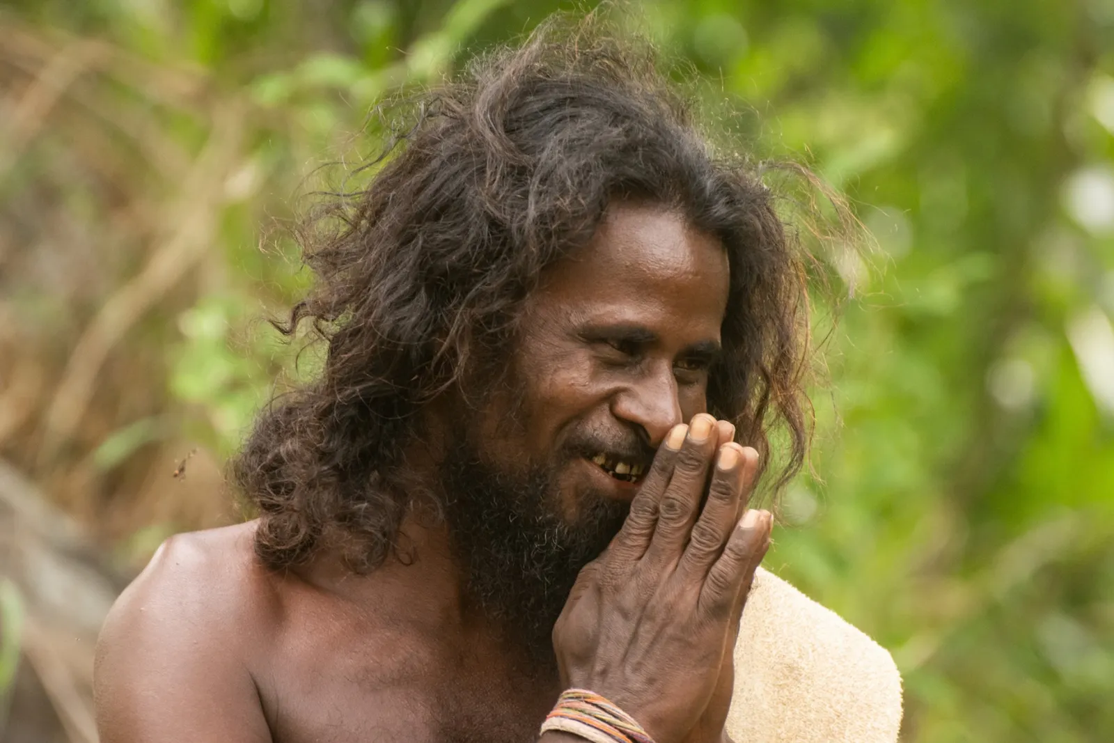 A long-haired elder with a beard, smiling with his hands pressed together, in front of forest greenery